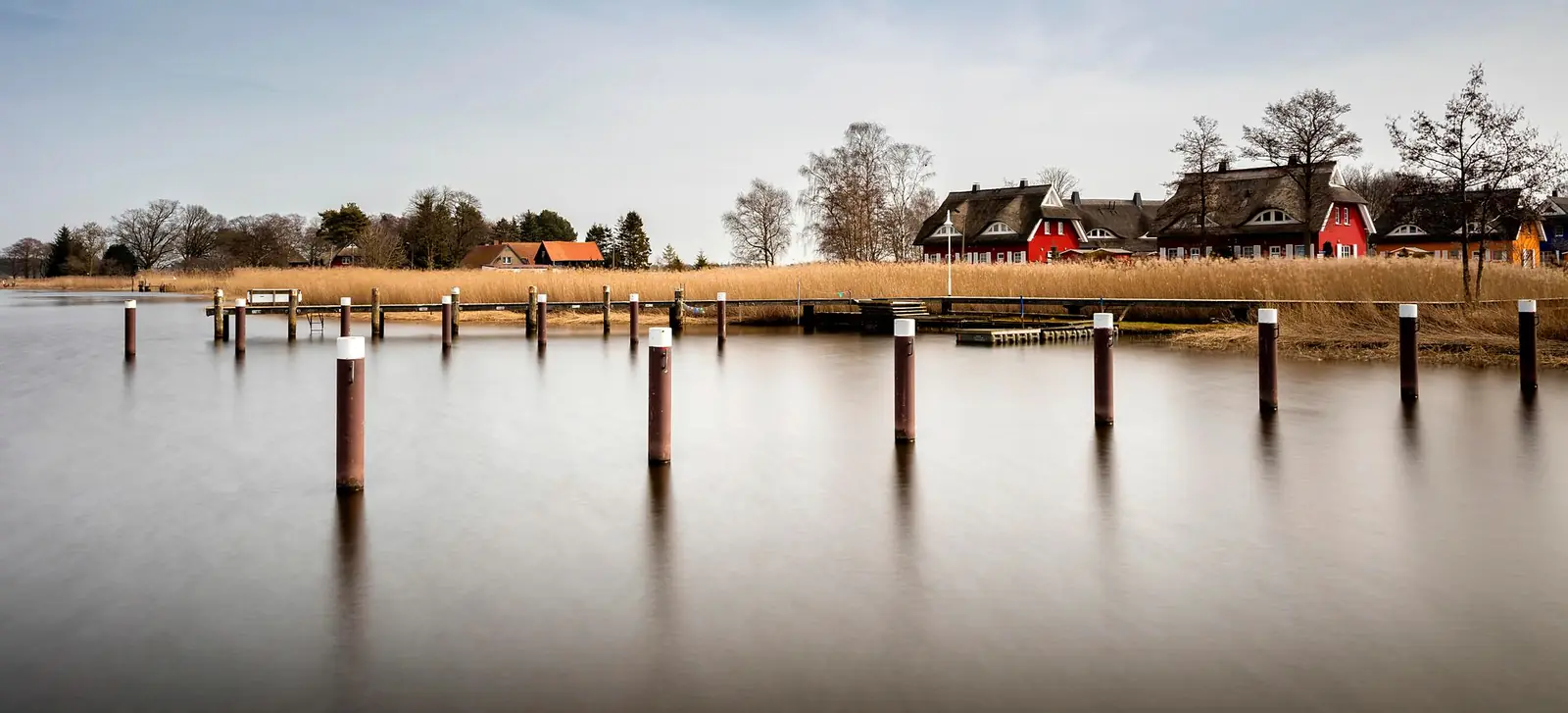 Dalben im spiegelglatten Wasser vor Schilf und Rohrdachhäusern